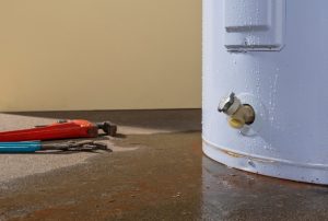 White water heater is leaking from the bottom, spilling water onto the floor next to a pair of repair tools.