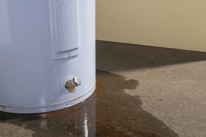 A white water heater leaking from the bottom, soaking the concrete floor around it.