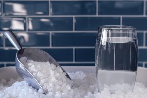 Glass of clear water beside water softener salt pellets and a metal scoop, representing water softening and hard water treatment.