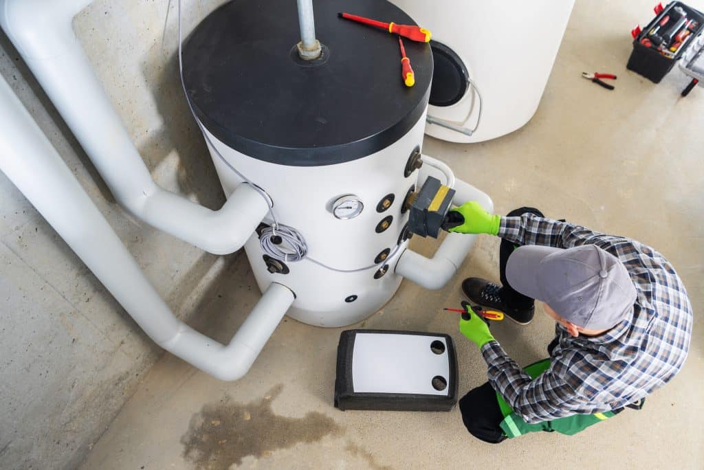 Plumber performing maintenance on a residential water heater tank, checking connections and controls with tools in a utility room.