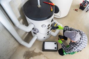 Plumber performing maintenance on a residential water heater tank, checking connections and controls with tools in a utility room.
