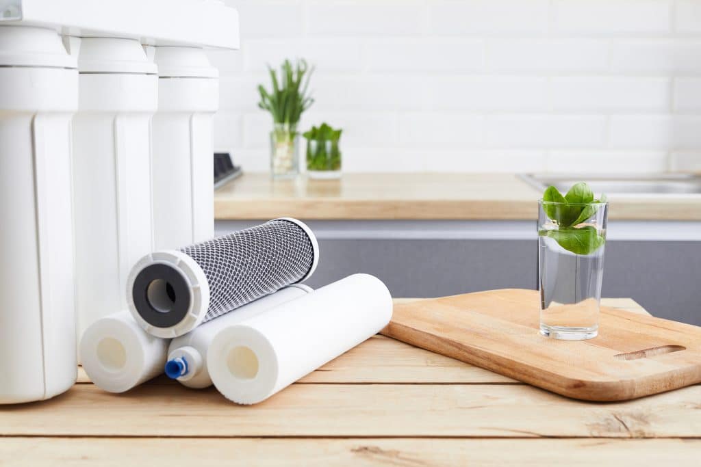 A glass of clean water with an osmosis filter and cartridges on a wooden table in a kitchen interior.